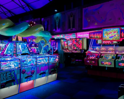 An indoor arcade filled with vibrant gaming machines lit with neon blue and pink lights. The scene includes various arcade games with flashing screens and colorful displays, and a large overhead structure resembling a green snake. The background shows illuminated signs and themed decor, contributing to an immersive entertainment atmosphere.