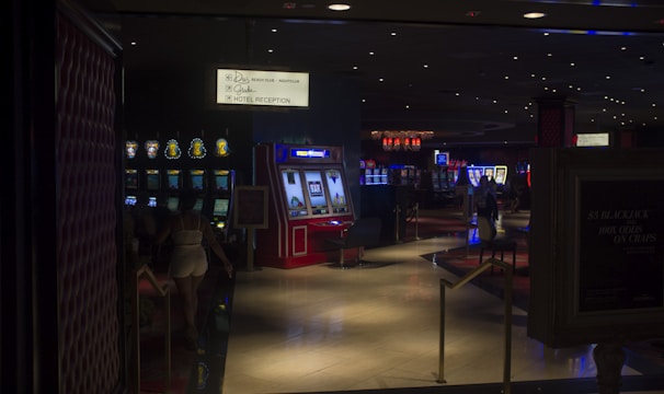 A dimly lit casino with several rows of slot machines, some displaying bright graphics and lights. There are a few people scattered throughout the area, engaging with the machines. The floor is reflective and polished, adding to the ambiance, while the ceiling is dotted with numerous small lights.