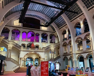 The interior of a large, ornately designed building with arched ceilings and intricate architectural details. The space features gaming machines lined up along the floor, under a high ceiling with a grid pattern. Lighting in purple and white tones enhances the ambiance.