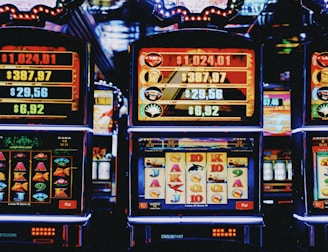 A row of colorful slot machines lit with bright neon lights, showcasing various graphical elements on the screens, indicating different games and jackpots. The atmosphere suggests a lively casino setting, bustling with excitement.