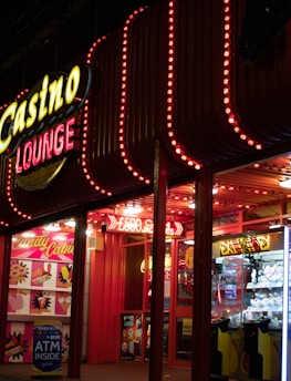 A brightly lit casino lounge entrance adorned with neon lights. The exterior features colorful signage including the words 'Casino' and 'Lounge' in vivid colors, with additional lights framing the building. Inside, a glimpse of a claw machine filled with plush toys and arcade games is visible. The area exudes an inviting yet bustling atmosphere typical of entertainment venues.