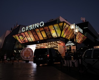 A building with a brightly illuminated entrance is seen at night. The sign above reads 'Casino' in bright letters. The facade features colorful graphics depicting various indulgent themes. Several vehicles are parked near the entrance, and people are visible in the vicinity. Tall lamp posts with spherical lights are present, casting a warm glow.